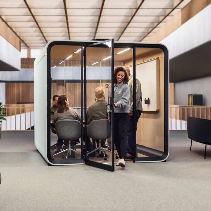 a soundproof booth at a commercial space with a person leaving the booth and 4 inside in a meeting