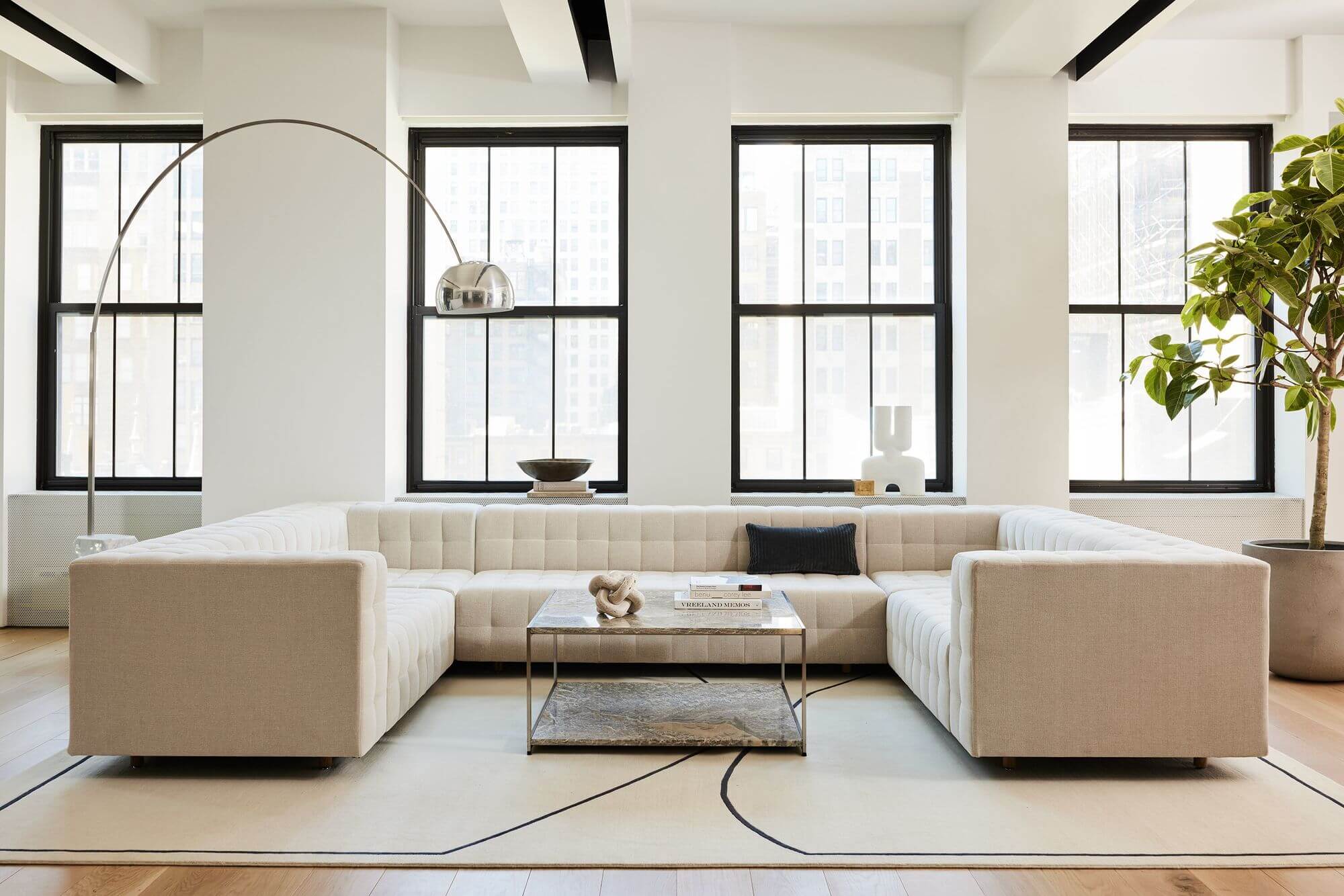 a U shaped Rapport Sofa in cream colour and a blue coushons in a white room and a middle coffee table with magazines in the middle with four windows in the background