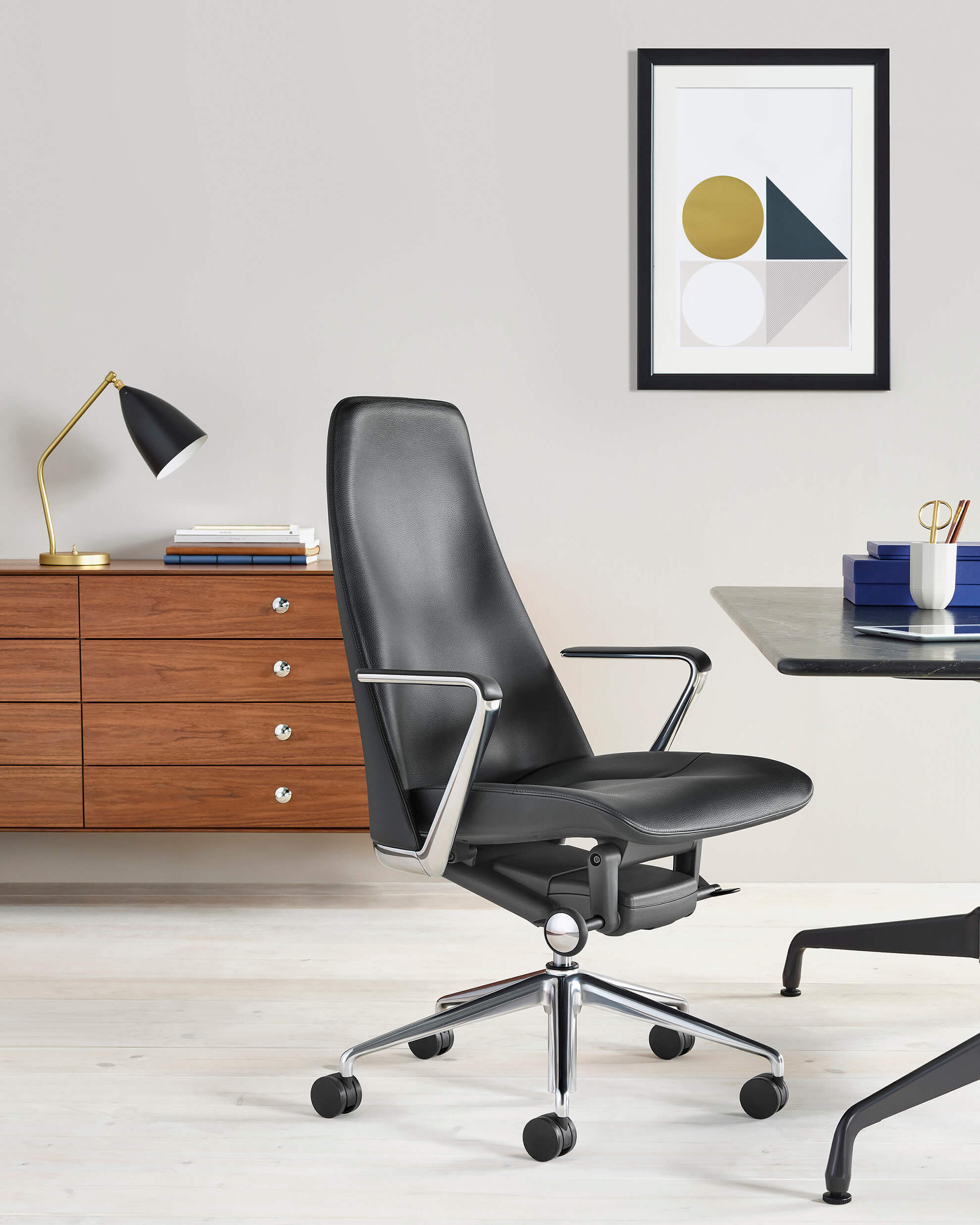 The Front Side angle of a Taper Executive Chair in black leather and polished aluminium frame with swivel base and castor wheels, in a modern office next to a desk and a storage cabinet behind with a painting in the top right