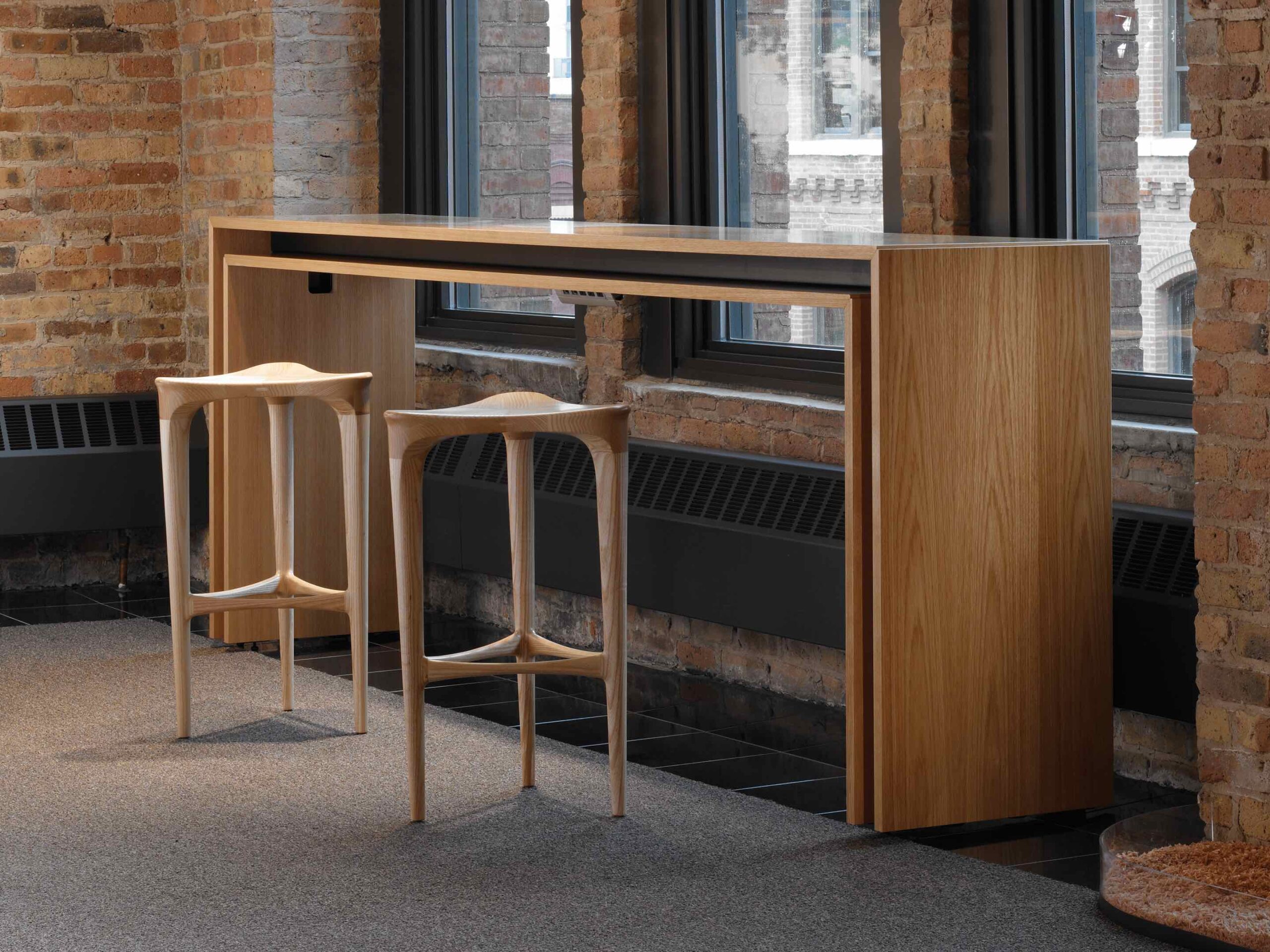 the side angle of a tall wooden white top Peer Conference Table featuring two wooden barstools on the sides and a brick wall with big windows in the background