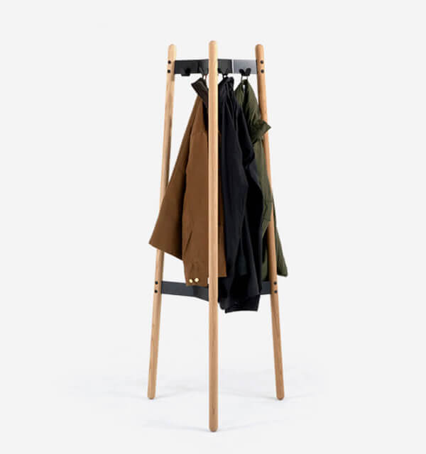 the fron side of a Hudson Coat Stand with wooden legs and metallic frame with 3 coats hanging on it in a white room