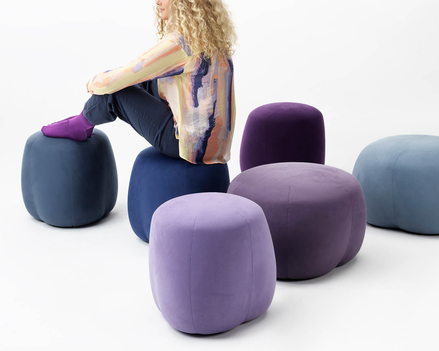 the close up if a person sitting on a Truffle Pouf blue colours, next to other Truffle poufs in a white room