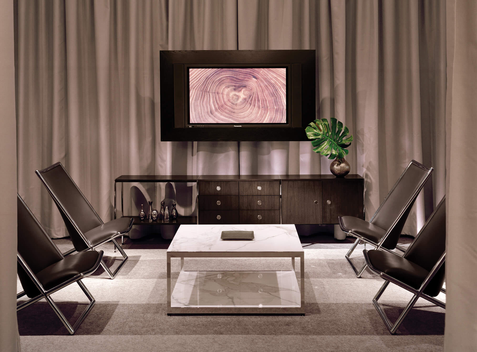 the front view of a two levels h frame storage cabinet with black wood top and drawers with polished aluminium legs in a lounge room featuring side leather and metalic frame chairs and a TV mounted in the background wall