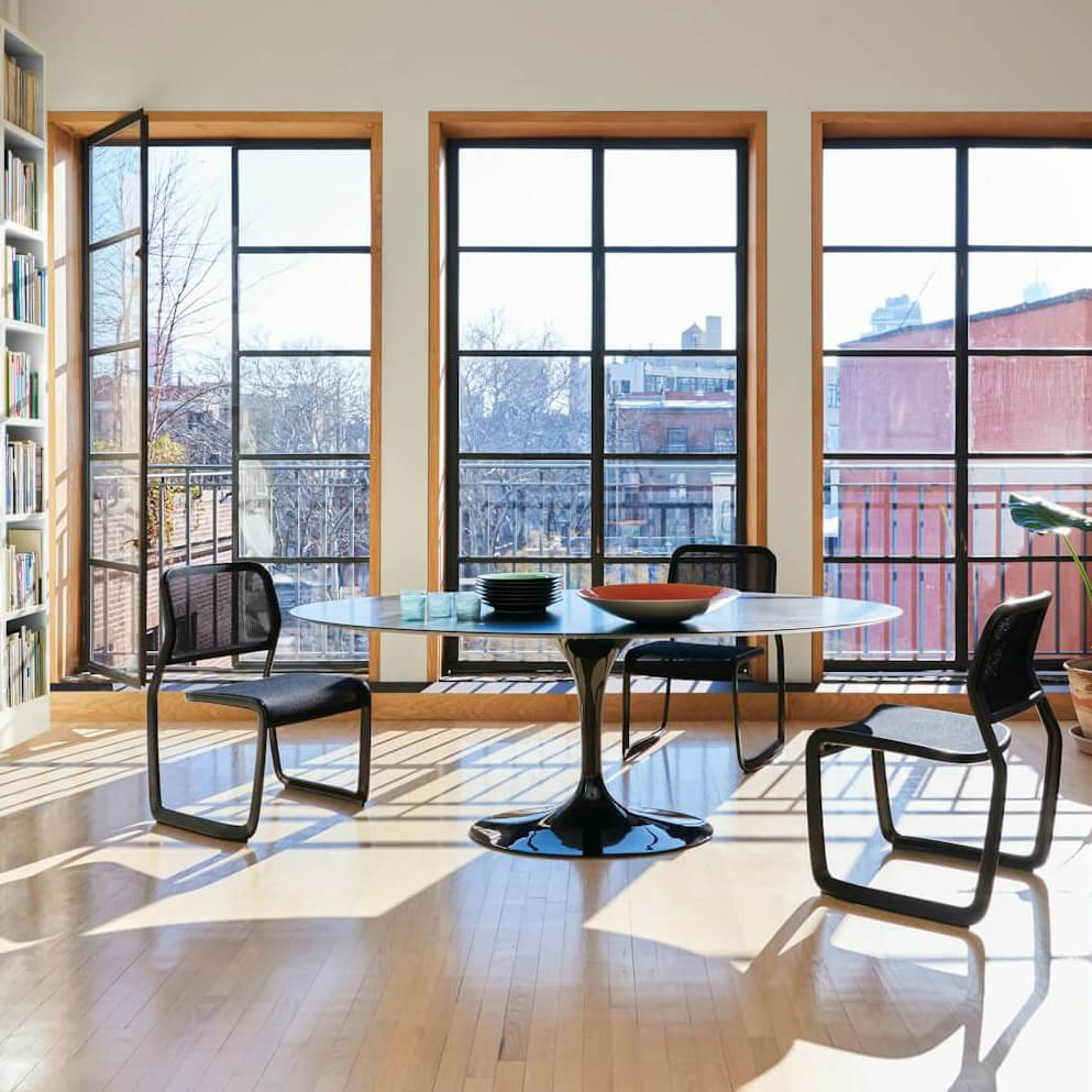 a modern library space featuring three tall windows in the background and a book shelf in the left side with a black rounded table in the middle surrounded by three Newson Aluminum Stacking Chairs