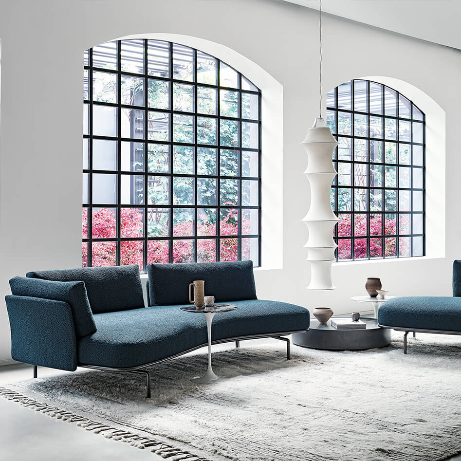 a minimalistic lounge area featuring two big wiondows in the backroung and a blue two seaters panoramic lounge sofa in the middle next to a side table and a light gray rugg