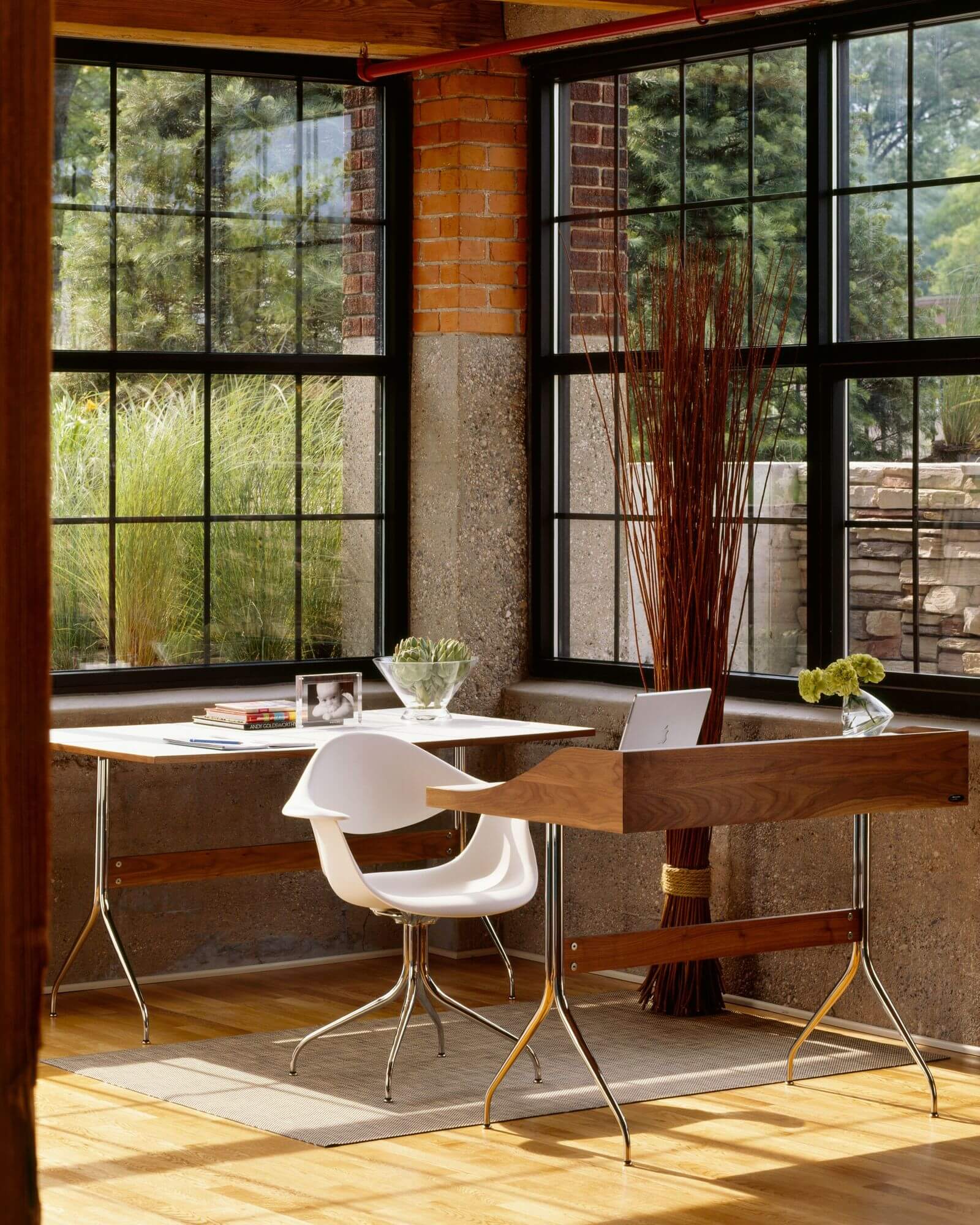 a mid century style home office studio featuring a Nelson Swag Leg Armchair and a wooden desk next to big windoes in a rustic home
